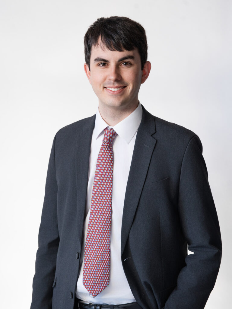 Professional headshot of a young man with short dark hair, wearing a dark gray suit, white dress shirt, and a red patterned tie. He is smiling confidently against a clean white background, with even lighting creating a polished and professional look.

