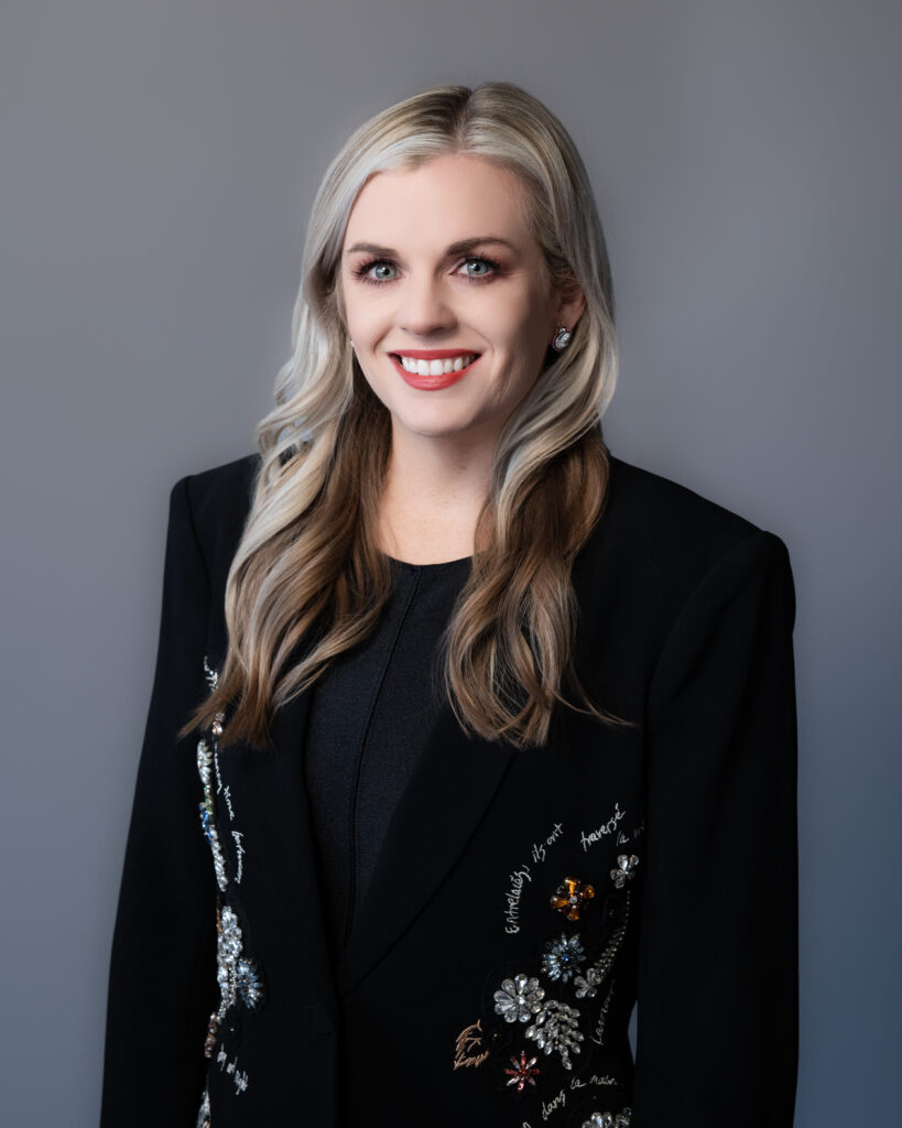 Professional headshot of a woman with long blonde hair, wearing a black blazer adorned with intricate embellishments and embroidery. She is smiling warmly against a neutral gray background, with soft and even lighting enhancing her polished and approachable look.